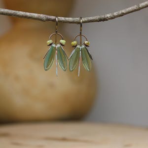 May include: A pair of handmade earrings with a nature-inspired design. The earrings feature green, leaf-shaped glass elements, copper accents, and small yellow beads. They hang from gold-colored ear wires, creating a delicate and artistic look.