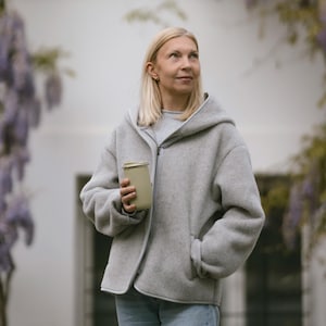 May include: A woman wearing a gray hooded jacket with a zipper and a pocket on the left side. She is holding a light green reusable coffee cup in her right hand.