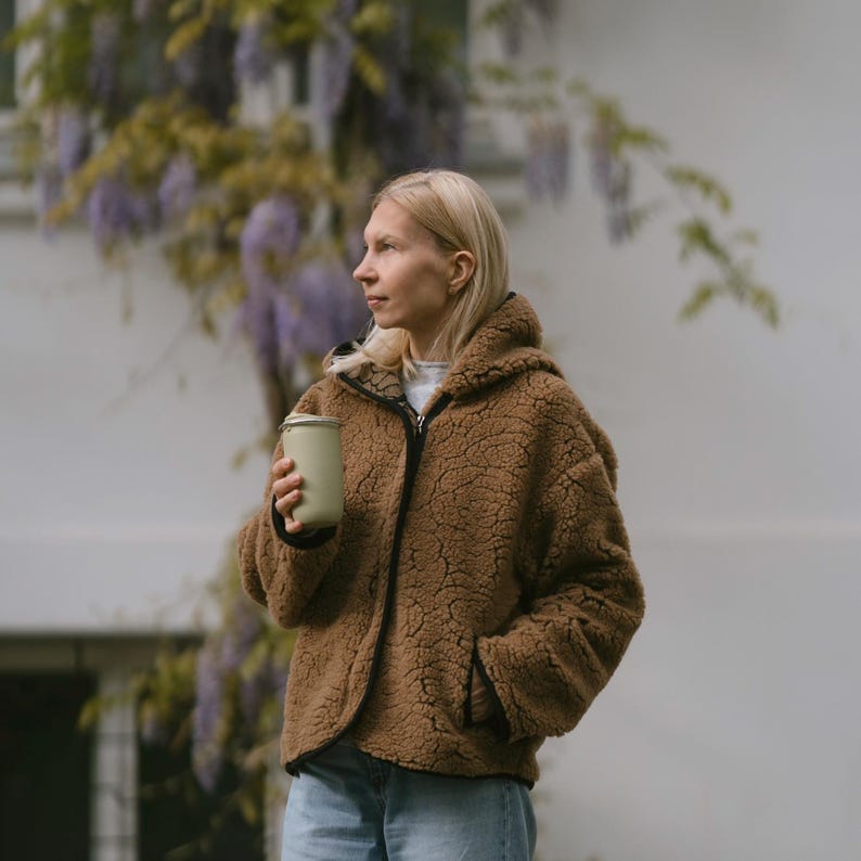 May include: A woman wearing a brown fuzzy hooded jacket holds a light green reusable cup. She is standing in front of a white wall with a purple flowering vine.