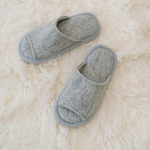 Peut inclure: Une paire de pantoufles en feutre gris &agrave; bout ouvert. Les pantoufles sont sur une surface blanche et moelleuse.