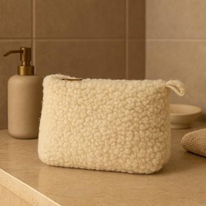 May include: Cream-colored, rectangular cosmetic bag with a fluffy, textured exterior. The bag has a small loop on the side and is sitting on a light-colored surface. A soap dispenser and other bathroom accessories are in the background.