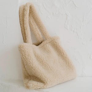 Puede incluir: Una bolsa de mano de color crema peluda con dos asas. La bolsa está hecha de un material suave y afelpado.