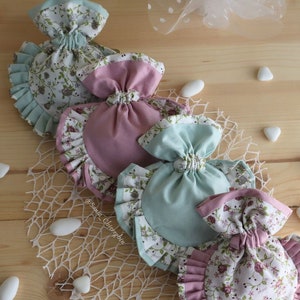 May include: Four fabric gift bags with floral patterns and ruffled edges. The bags are in shades of pink, green, and white. The bags are arranged in a row on a wooden surface.