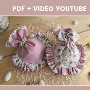 May include: Two small pink and white fabric pouches with floral print and ruffled edges. The pouches are tied with a bow and are sitting on a wooden surface with white stones.