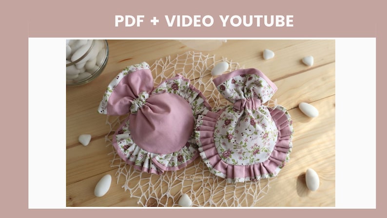 May include: Two small pink and white fabric pouches with floral print and ruffled edges. The pouches are tied with a bow and are sitting on a wooden surface with white stones.