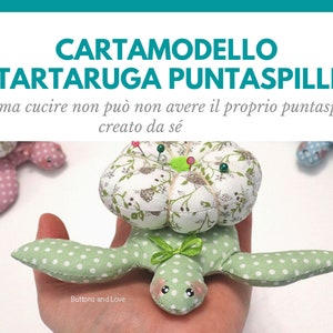 Può includere: Un cuscino per spilli a forma di tartaruga a pois verdi e bianchi con un top a stampa floreale. Il cuscino per spilli è pieno di spilli e si trova su una mano. Il testo "CARTAMODELLO TARTARUGA PUNTASPILLI" è in alto nell'immagine. Il testo "chi ama cucire non può non avere il proprio puntaspilli creato da sé" è sotto la tartaruga. Il testo "Buttons and Love" è in basso nell'immagine.
