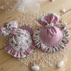 May include: Two pink fabric pouches with ruffled edges. One pouch is decorated with a floral pattern and the other is solid pink. Both pouches are sitting on a white crocheted doily.