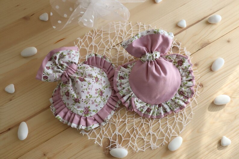 May include: Two pink fabric pouches with ruffled edges. One pouch is decorated with a floral pattern and the other is solid pink. Both pouches are sitting on a white crocheted doily.