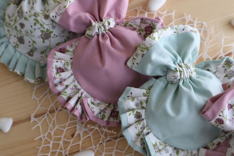 May include: Two fabric pouches with floral print and ruffled edges. One pouch is pink and the other is light blue. Both pouches are tied with a bow at the top.