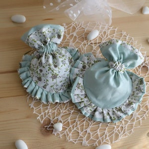 May include: Two small fabric bags with ruffled edges. One bag is light blue with a white floral pattern. The other bag is white with a light blue floral pattern. Both bags are tied with a bow.
