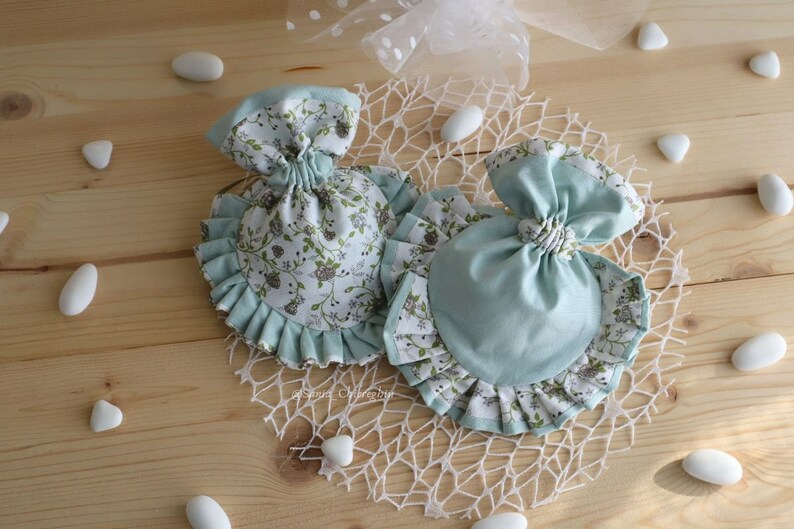 May include: Two small fabric bags with ruffled edges. One bag is light blue with a white floral pattern. The other bag is white with a light blue floral pattern. Both bags are tied with a bow.