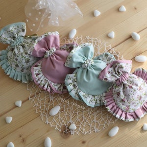 May include: Four fabric gift bags with floral patterns and ruffled edges. Two bags are pink and two are light blue. The bags are arranged in a circle on a wooden surface with white stones scattered around them. The bags have a bow at the top and are tied with a ribbon.