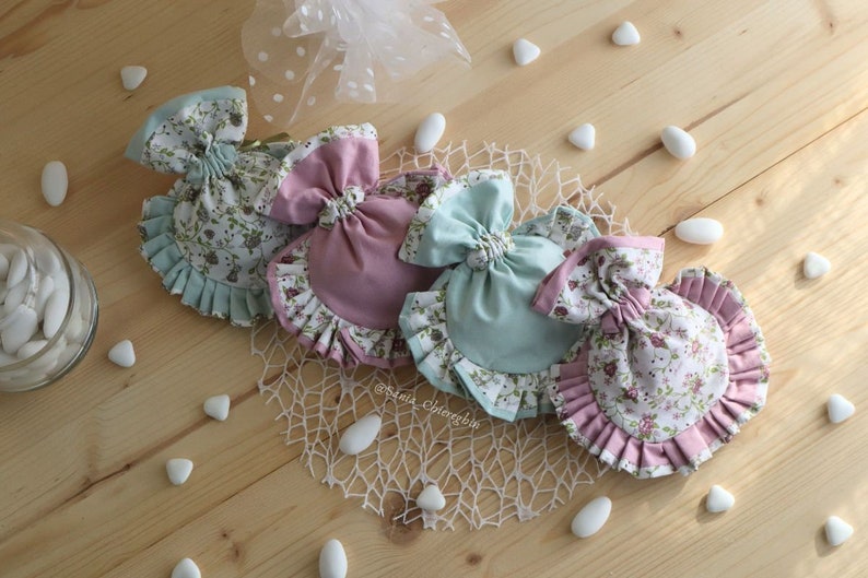 May include: Four fabric gift bags with floral patterns and ruffled edges. Two bags are pink and two are light blue. The bags are arranged in a circle on a wooden surface with white stones scattered around them. The bags have a bow at the top and are tied with a ribbon.