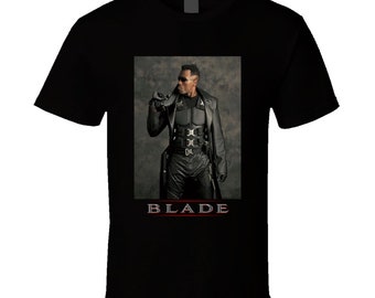 May include: Black t-shirt featuring a centered image of a man in a leather outfit and sunglasses. The image includes the word "BLADE" in red and white text at the bottom. The man is holding a pair of sunglasses.