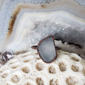 May include: A handcrafted ring featuring a piece of sea glass in a light blue-gray hue, set in a copper-colored, textured metal band. The sea glass has a unique, irregular shape. The ring is displayed on a white, textured surface.