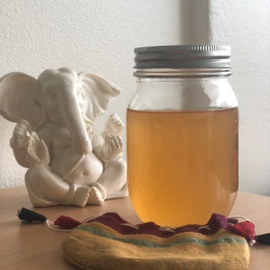 Pode incluir: Um frasco de vidro transparente com uma tampa prateada cheio de um líquido âmbar claro. O frasco está sentado em uma superfície de madeira ao lado de uma pequena figura de elefante branco e uma bolsa de tecido amarela e vermelha com uma borla preta.