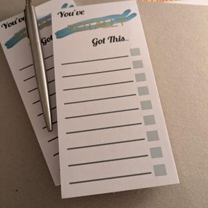 Puede incluir: Dos blocs de notas blancos con el texto "You've TOTALLY Got This..." en letras negras y doradas, con un diseño de pincelada azul. Un bolígrafo plateado descansa sobre los blocs de notas. Los blocs de notas tienen líneas y casillas de verificación.
