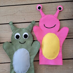 May include: Two felt hand puppets, one green with gray eyes and a smiling mouth, the other pink with yellow eyes and a smiling mouth.