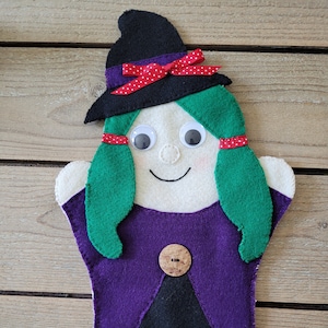 May include: A felt witch puppet with a black hat, green hair, purple dress, and a brown button. The puppet has white eyes and a smiling mouth.