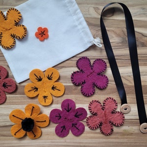 May include: A set of felt flower shapes in orange, yellow, pink, and red with black stitching. The flowers are arranged on a wooden surface with a black ribbon and two wooden buttons.