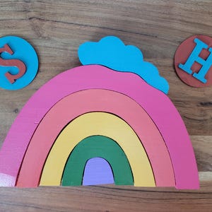 May include: A rainbow-colored wooden arch with a blue cloud on top. The arch is made of six pieces of wood, each painted a different color of the rainbow. The arch is sitting on a wooden surface. There are two wooden circles with the letters "S" and "H" on them.