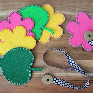 May include: A collection of colorful felt flower and leaf shapes. The flowers are pink and yellow, with a green leaf. The edges are stitched with contrasting thread. A pink flower has a button and a black and white polka dot ribbon.