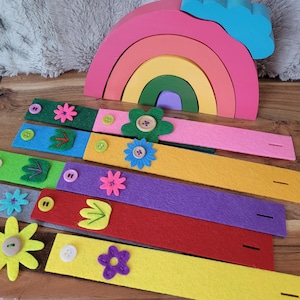 May include: A set of colorful felt strips with various flower and leaf designs, each with a button and a slit. The strips are arranged in a rainbow pattern.