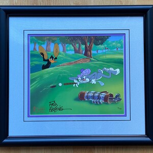 Puede incluir: Una ilustración de dibujos animados enmarcada de Bugs Bunny y el Pato Lucas jugando al golf. Bugs Bunny está tumbado en el suelo, balanceando un palo de golf, mientras que el Pato Lucas está de pie detrás de él, sosteniendo dos palos de golf. La imagen está firmada "For the Fans" y tiene un logotipo de Warner Bros.