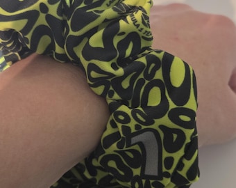 Peut inclure: Un chouchou avec un motif audacieux de formes noires sur un fond jaune fluo. Le chouchou est porté autour d'un poignet. Le texte "LANDO Norris" et "F1" sont imprimés sur le tissu.