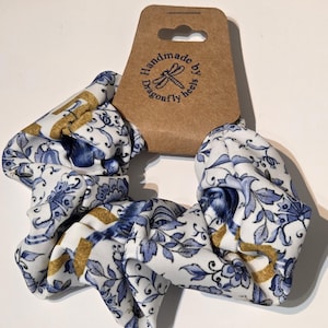 May include: A handmade scrunchie featuring a white base with a blue floral design and gold stripes. The scrunchie is attached to a brown tag with the text "Handmade by dragonfly heels."
