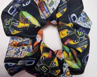 Peut inclure: Un chouchou noir avec un motif répété de graphiques de voitures de course colorées, de logos et de texte. Le chouchou présente une variété de couleurs, dont le jaune, le bleu, le rouge et le vert. Le tissu est froncé et torsadé.