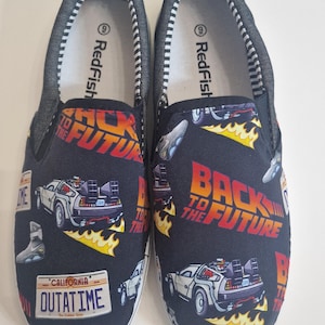 Può includere: Un paio di scarpe da barca in tela nera con un motivo ripetuto della DeLorean, la macchina del tempo del film Ritorno al futuro. Le scarpe hanno una suola bianca e una fodera interna a righe bianche e nere. Il testo "Redfish" è stampato sulla fodera interna di ogni scarpa.
