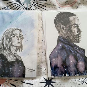 Puede incluir: Dos impresiones artísticas con retratos. Una muestra a una mujer con cabello largo sobre un fondo azul. La otra representa a un hombre de perfil, con una chaqueta con temática de galaxia. Ambas son en blanco y negro con detalles en acuarela.