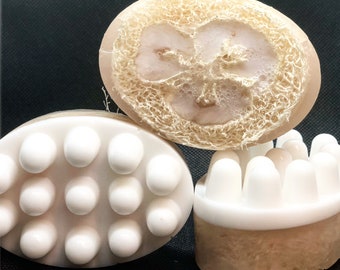 May include: Three handmade soaps are displayed. One is oval-shaped with white massage nodules, another is round with a loofah center, and the third is rectangular with white massage nodules. The soaps are light brown and white.