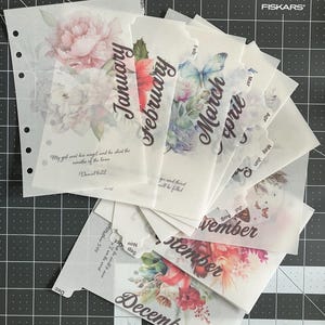 May include: Twelve monthly planner dividers featuring floral designs and the month name in cursive script. Each divider includes a quote or scripture verse printed on it. The dividers are made from clear plastic and are designed to fit a standard planner.
