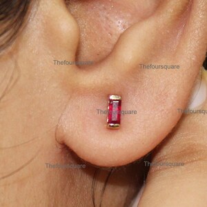 May include: A close-up of a person's ear with a small gold earring featuring a red rectangular gemstone.