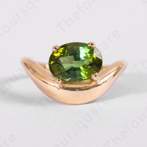 May include: A gold ring featuring a vibrant green oval gemstone. The ring's band has a unique crescent shape, with the gemstone securely held in place by four prongs. The ring is set against a white background.