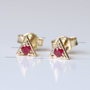 May include: A pair of gold triangle stud earrings with a red gemstone in the center of each triangle.
