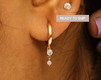 May include: Gold hoop earrings with a dangling chain and three small clear stones. The earrings are shown on a person's ear.