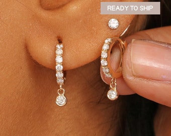 May include: A pair of gold hoop earrings with a dangling diamond. The hoops are set with small diamonds and the dangling diamond is larger. The earrings are shown on a person's ear.