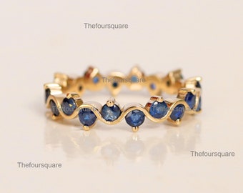 May include: A gold-colored ring featuring a continuous row of round, deep blue gemstones. The gemstones are evenly spaced and set within a wavy, bezel-style design. The ring is displayed on a light-colored surface.