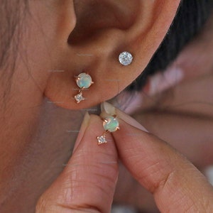 May include: A pair of dainty earrings featuring a small round opal gemstone with a tiny diamond accent. The earrings are made of rose gold and have a minimalist design.