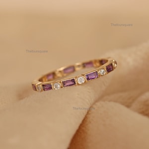May include: A gold band ring with a row of alternating purple and clear gemstones. The ring is on a light brown fabric background.