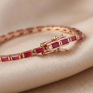 Könnte beinhalten: Ein goldfarbenes Armband mit einer durchgehenden Reihe rechteckiger, tiefroter Edelsteine in einer Metallfassung. Das Schmuckstück verfügt über einen sicheren Kastenschloss-Verschluss, der farblich auf die Metallglieder abgestimmt ist.