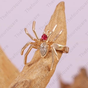 May include: A gold spider ring with a red gemstone and a clear diamond. The ring is set on a piece of driftwood.