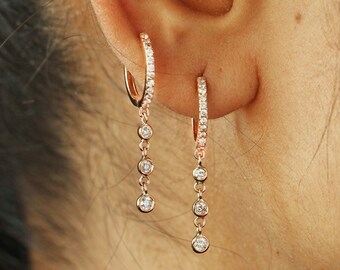 May include: A pair of rose gold hoop earrings with dangling charms. Each hoop is lined with small clear stones and has a drop of three clear stones.