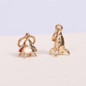 Puede incluir: Dos abalorios de color dorado, uno con forma de cohete y el otro con forma de OVNI. Ambos abalorios tienen un diseño detallado y son perfectos para añadir un toque de fantasía a las joyas.