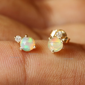 May include: A pair of gold stud earrings featuring a small round opal gemstone and a tiny diamond accent. The opal displays iridescent hues of green, blue, and pink.