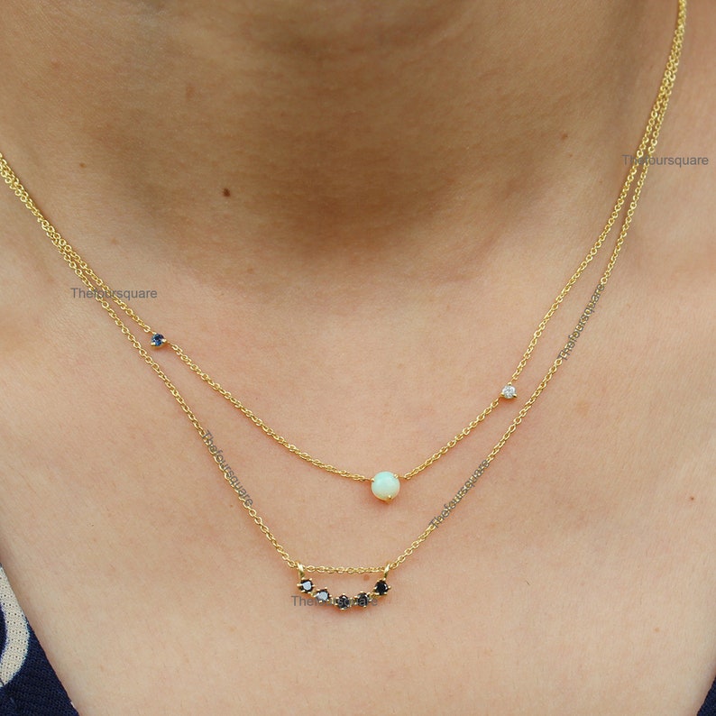 Op de afbeelding: Een gouden ketting met een witte opaal hanger en een rij zwarte edelstenen. De ketting is gelaagd met een tweede gouden ketting met een enkele witte edelsteen.