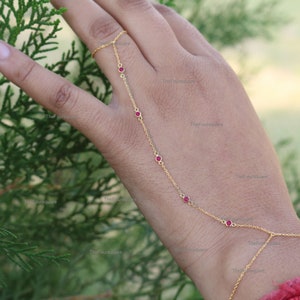 May include: A gold chain bracelet with red gemstones. The bracelet is designed to be worn on the finger and wrist.